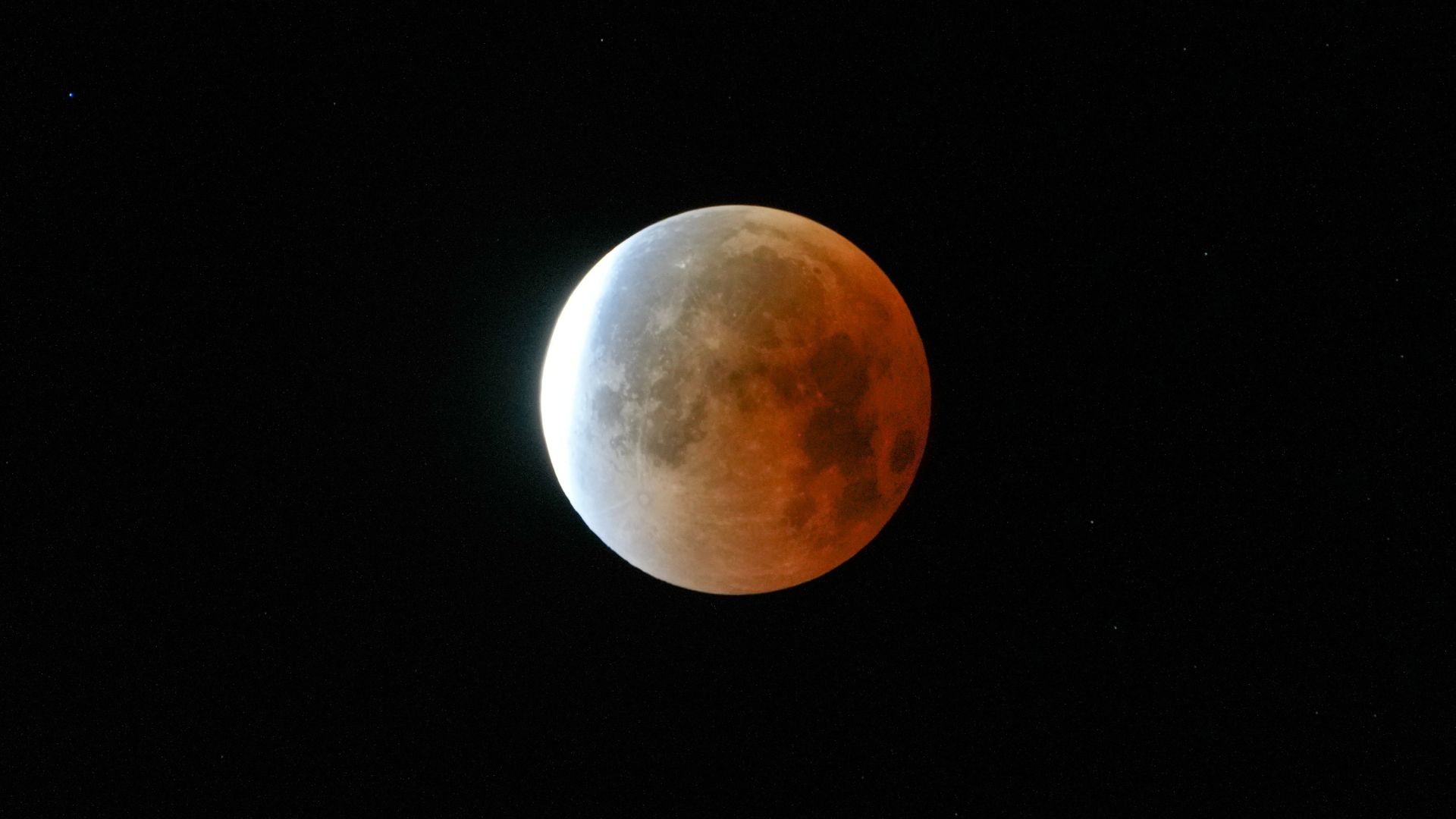A full moon is shown in a black sky during a lunar eclipse, with its right side glowing red in Earth's shadow.