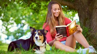 Young woman reading a book while sitting with her dog under a tree
