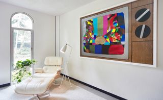 White armchair with footstool and standard lamp next to a brightly coloured painting on the wall
