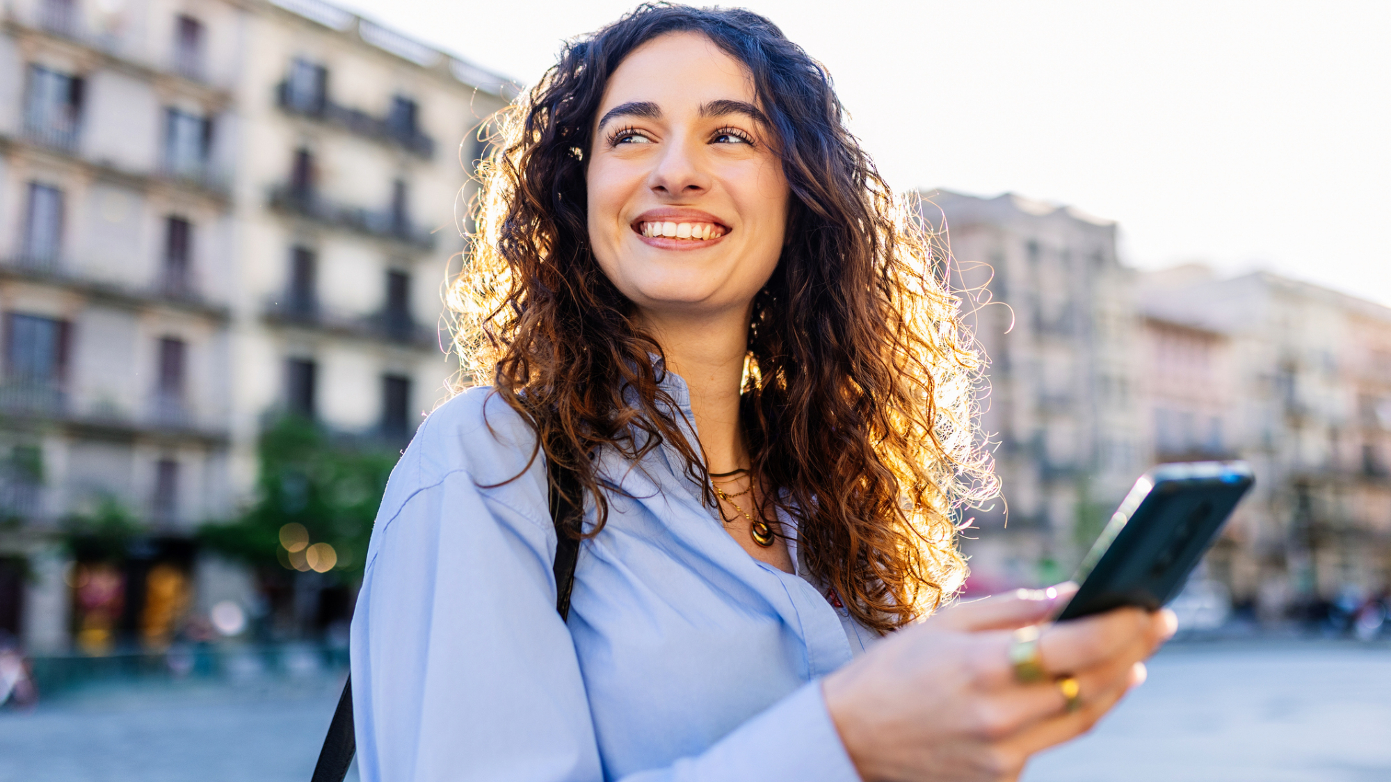 A woman, holding their phone, stood in front of buildings.