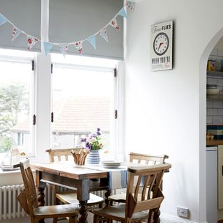 A white dining room with grey blinds, bunting and a small wooden table and chairs