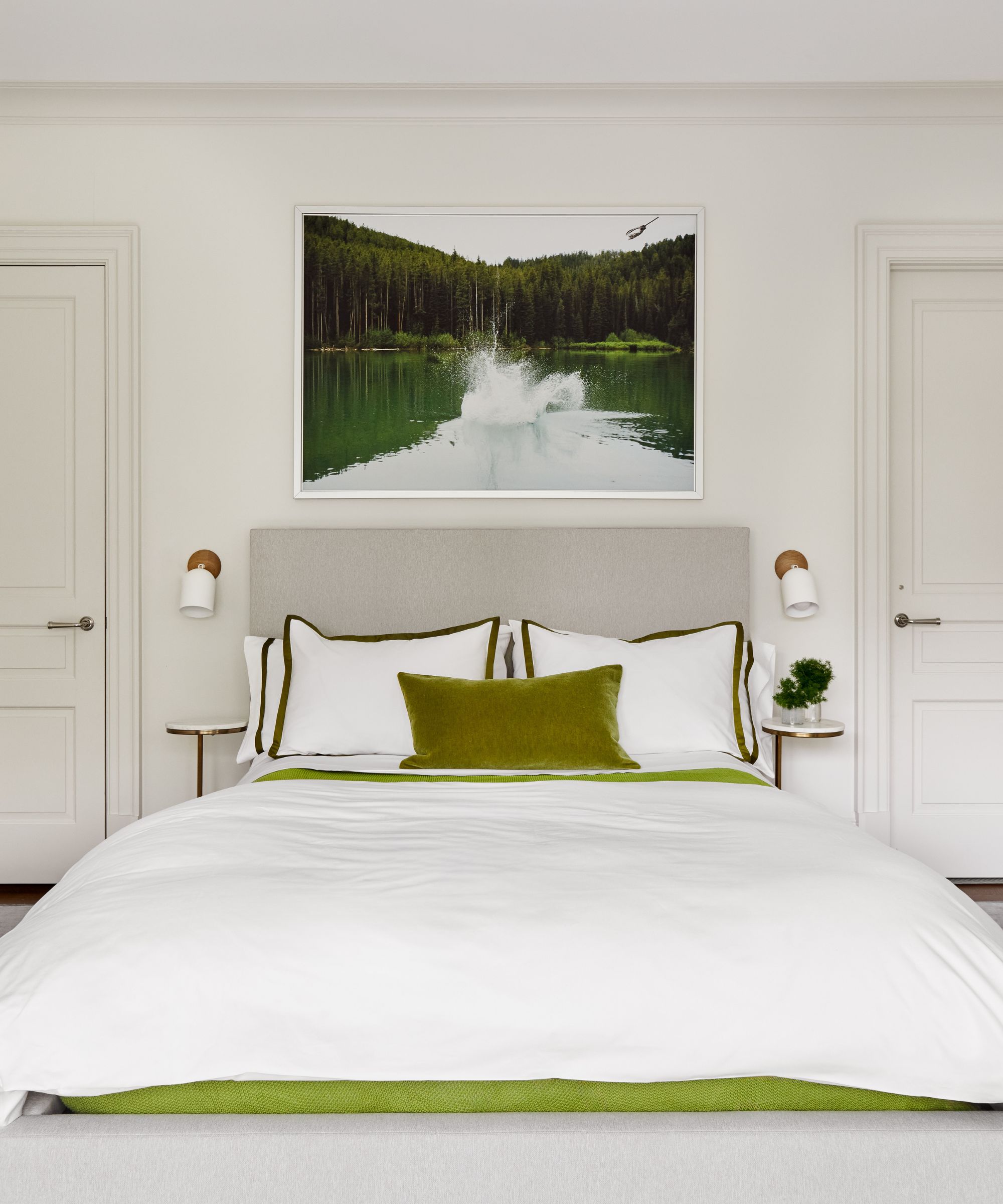 A bedroom with pale gray walls, a double bed with white bedding and moss green accents, and a green landscape picture on the wall.