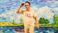 A man in light-colored clothing and a graphic tee stands inside a painted boat, posing with his hand to his forehead against a vibrant, impressionistic lake backdrop.