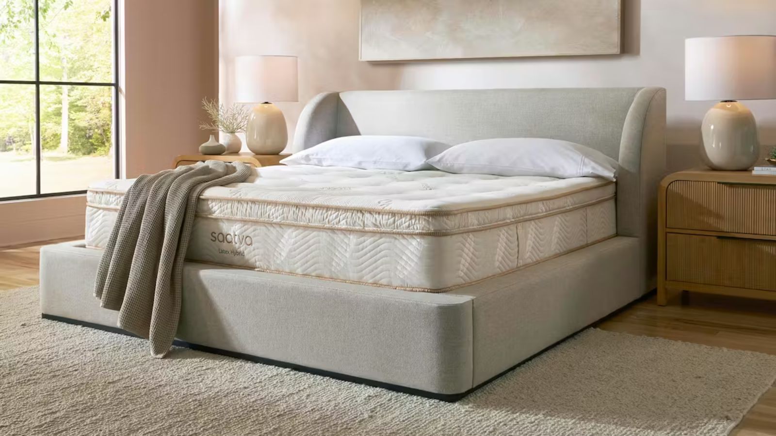 The Saatva Latex Hybrid Mattress on a grey boucle bed frame. To either side of the bed are matching wooden bedside tables with cream ceramic lamps with white lampshades, and beneath the bed is a grey rug. The walls are pale pink, and to the left is a large crittal window.