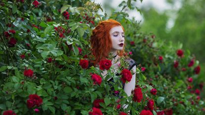 Young woman dressed in goth style stands amid a red rose bush