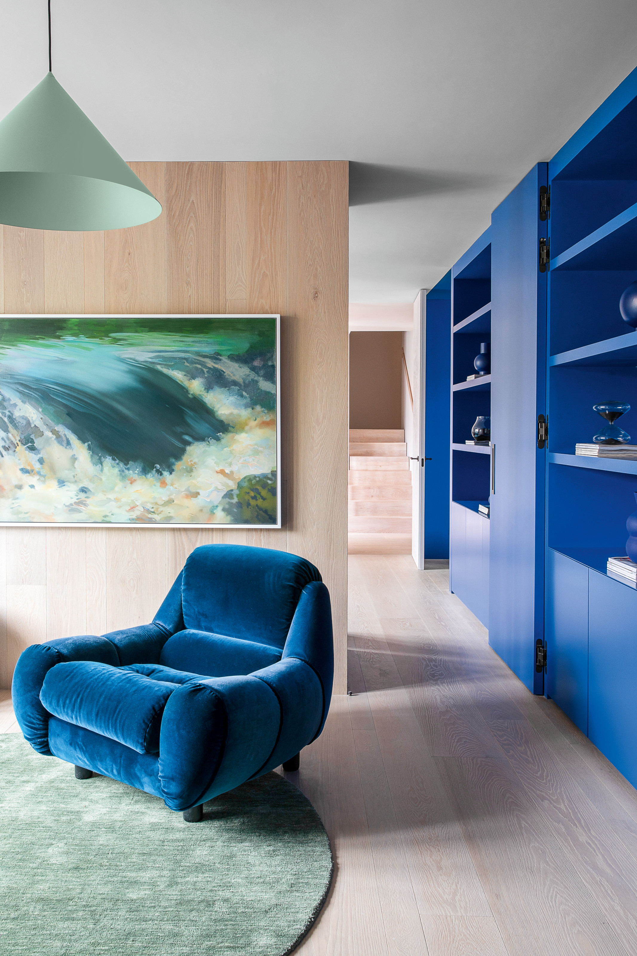 Room with pale wood cladding, electric blue painted shelving, electric blue armchair and aqua green rug, lamp shade and artwork