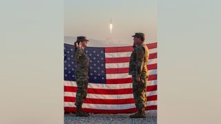 two people in combat fatigues salute one another in front of an American flag while a rocket launches above a plume of fire in the background