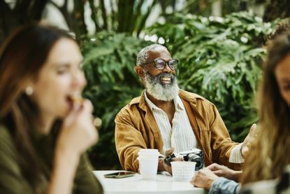 Medium shot of smiling senior man relaxing at outdoor caf&eacute; during vacation in city. He holds a camera.