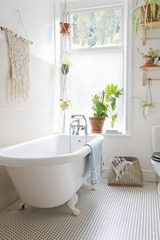 White bathroom with wall and floor mosaic tiles, white freestanding bath, plants on open shelving
