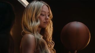 Sydney Sweeney sitting on a basketball hoop holding a basketball. She is wearing her hair.