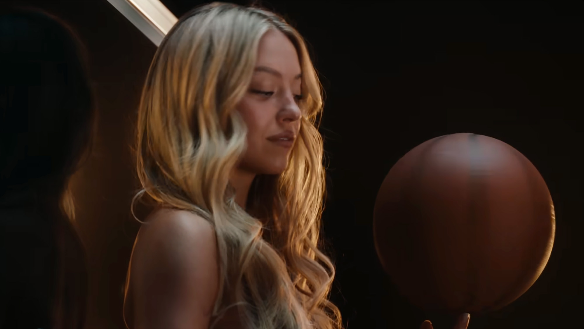 Sydney Sweeney sitting on a basketball hoop holding a basketball. She is wearing her hair.