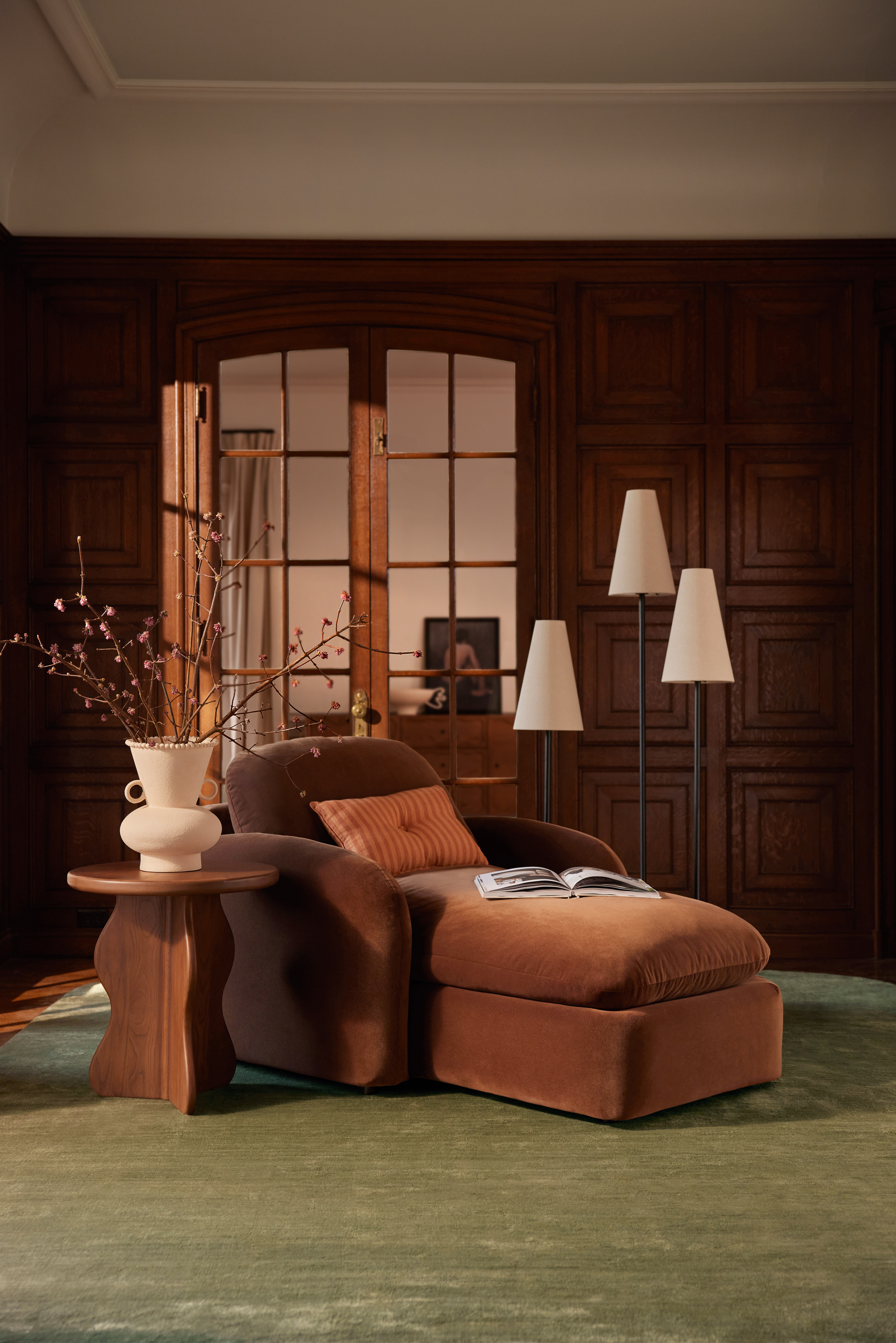 Lulu and Georgia scalloped wooden side table, velvet chaise lounge, and three-shaded floor lamp pixutred in a wood paneled living room with moss green carpet