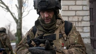 A Ukrainian military member holds a smartphone, with a residential building in the background. They are based in Donetsk Oblast, Ukraine.