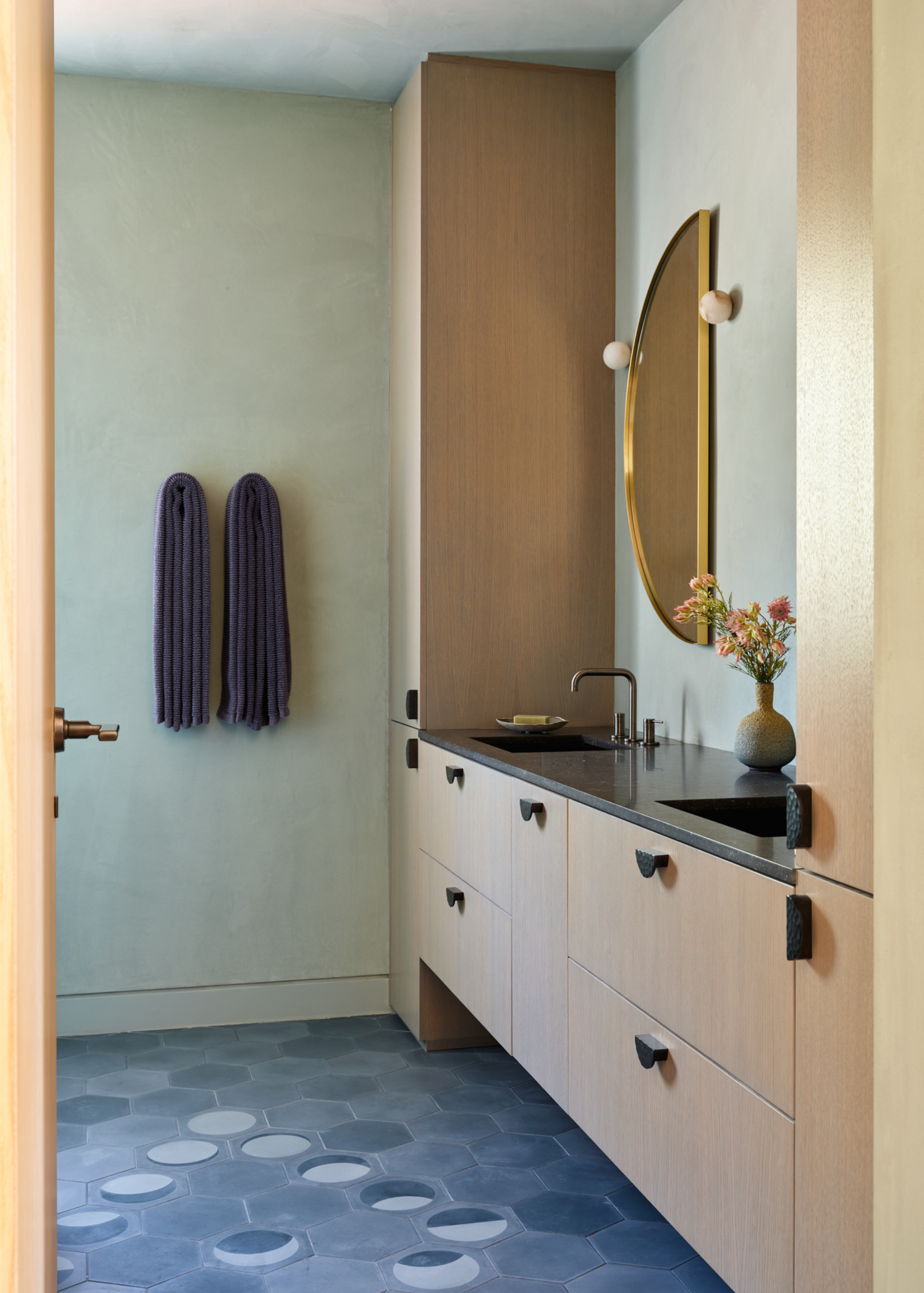A bathroom with dusty blue walls, a set of blue towels, wooden drawers and cabinets, a semi-circle mirror. wall sconces, and a vase of flowers