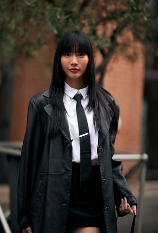 A woman wearing geek chic trend in the form of a leather jacket layered over a white shirt styled with a black tie and a black miniskirt during Milan Fashion Week spring/summer 2025.