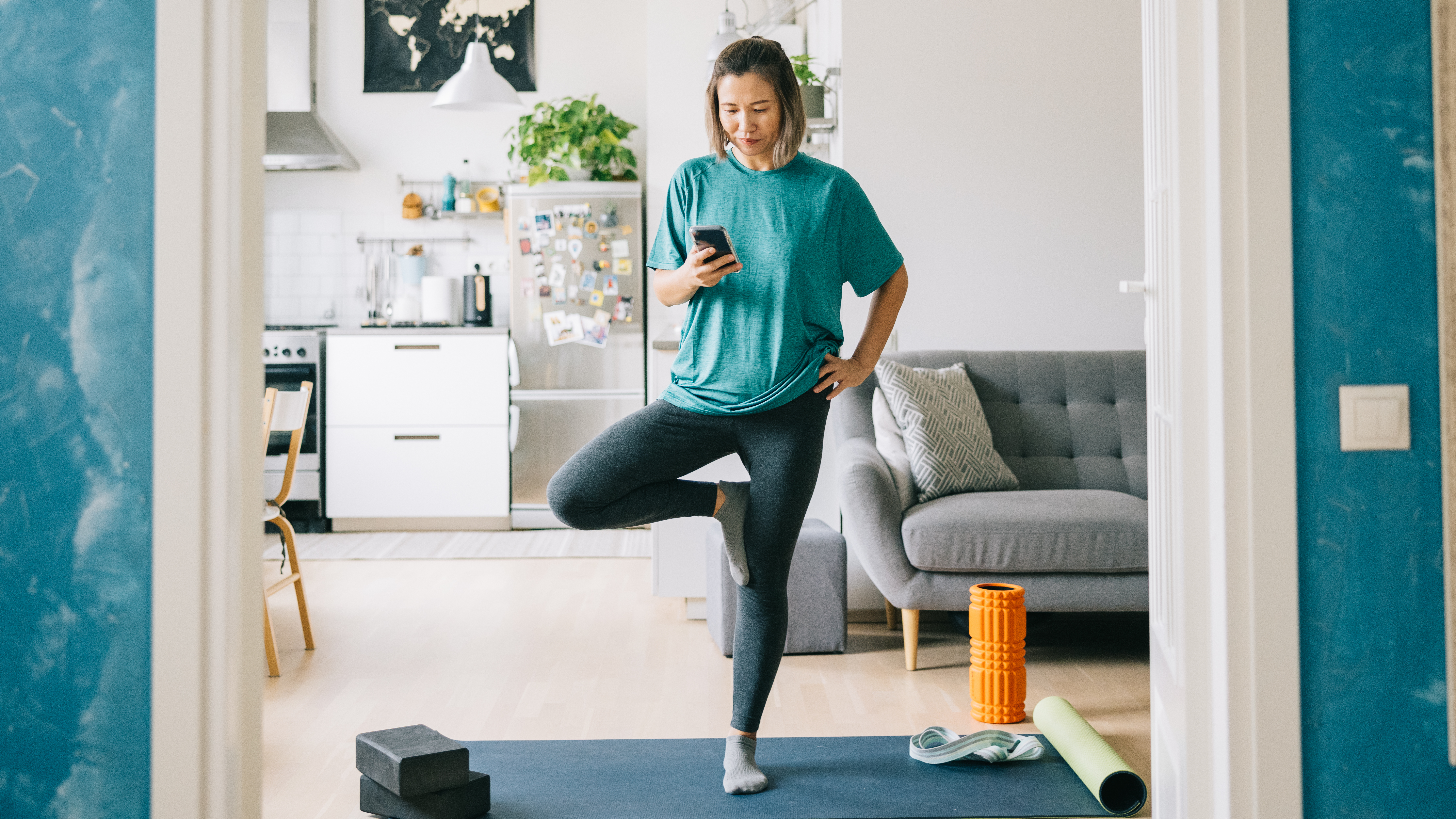 A woman stands on one leg on an exercise mat looking at her phone. Sh's in a living room/ kitchen area, with plants and pictures behind her.