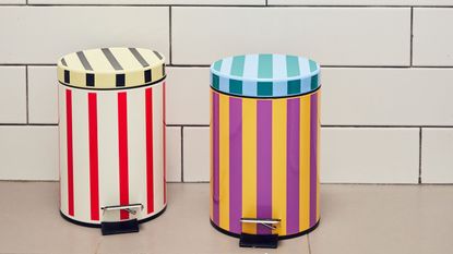 Two colorful and striped trash cans in a bathroom with a whit tiled backdrop. 