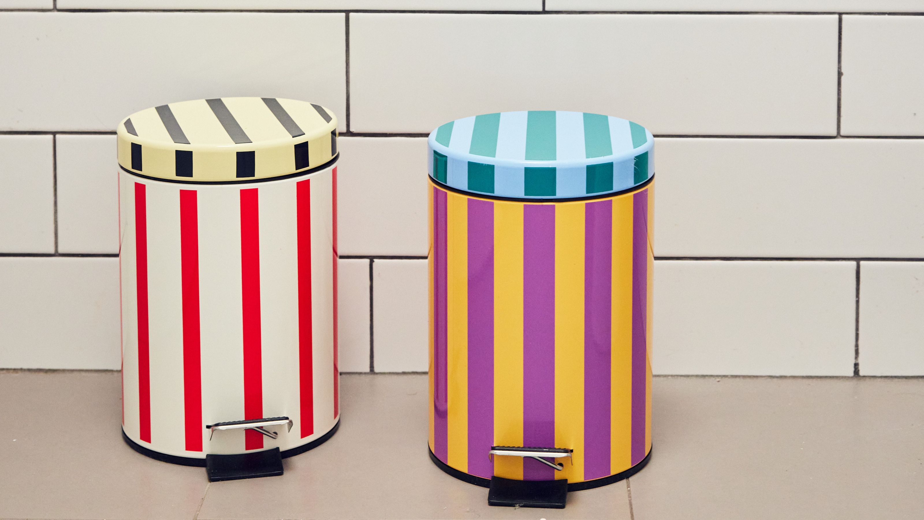 Two colorful and striped trash cans in a bathroom with a white tiled backdrop.