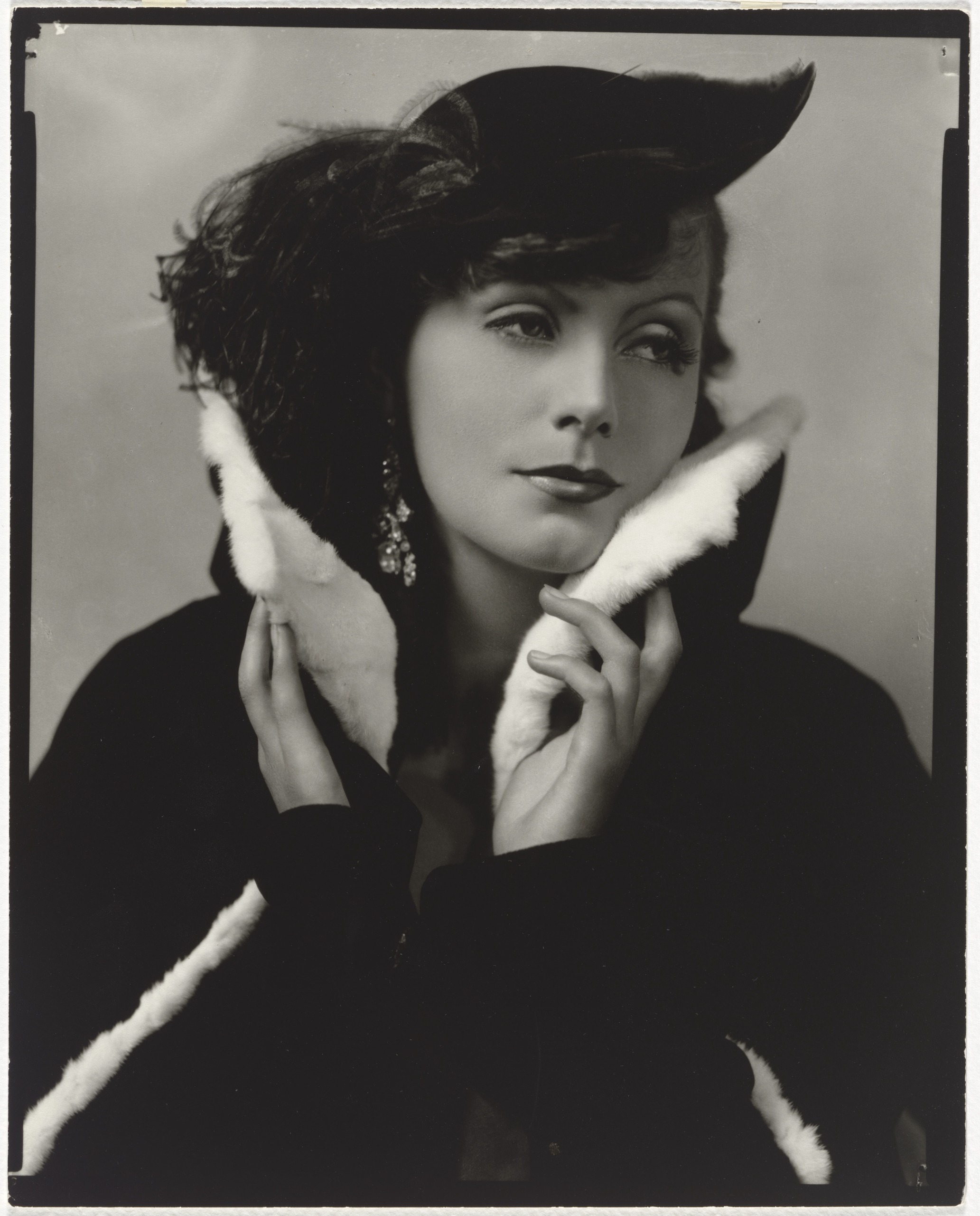 Greta Garbo in a dark coat with a white fur collar and a feathered hat holds the collar up to her face with both hands, gazing off to one side, with drop earrings catching the light.