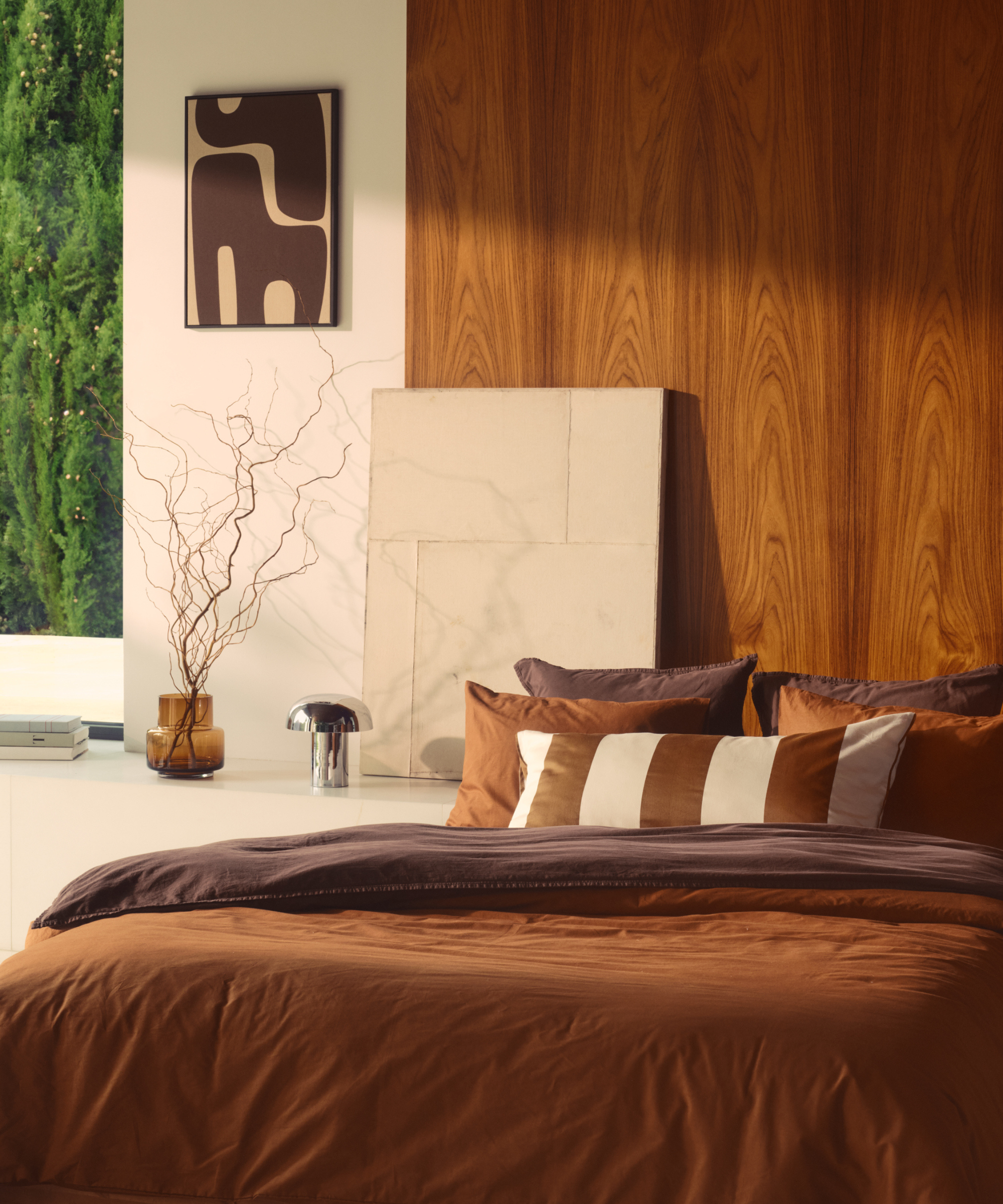 a bed with brown bedding, several pillows in shades of brown, a chrome table lamp, and a vase holding sticks