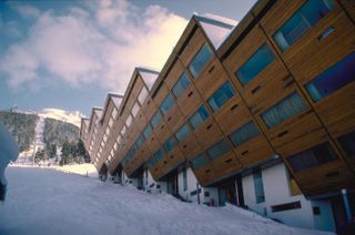 Exterior view of the hotel in a ski resort. To the right, a three-story Wooden building seems like it's leaning towards the ground and each section on the other. To the left, we see a skiing path.