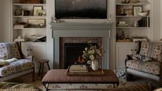 a neutral small living room in a carriage house with a soft green fireplace with expose brick detail, built in bookcases, floral armchairs, a striped ottoman and a woven rug with floral detail