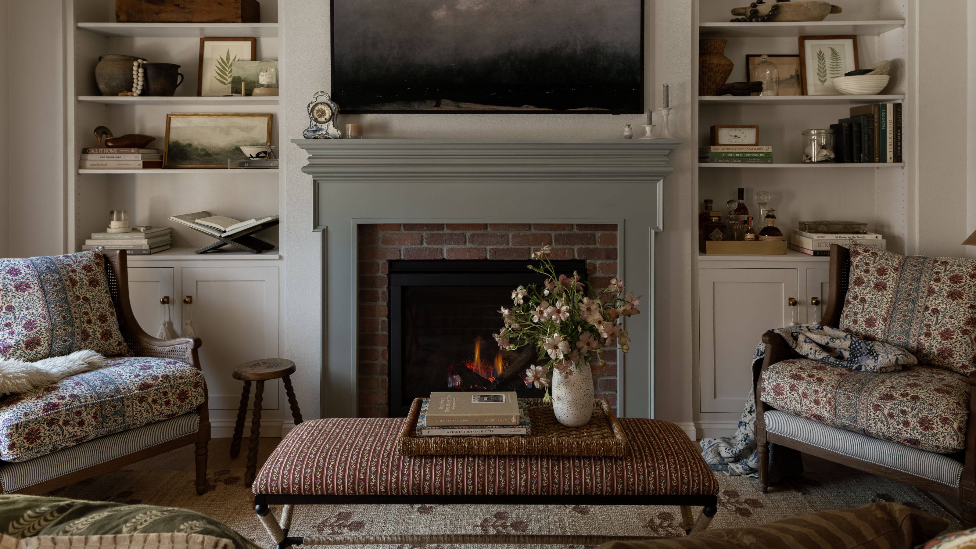a neutral small living room in a carriage house with a soft green fireplace with exposed brick detail, built in bookcases, floral armchairs, a striped ottoman and a woven rug with floral detail