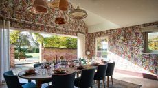 Image of an open room that leads to a back garden and has high slanted ceilings. There is a pink, floral-patterned wallpaper on the walls, and a dining table in the middle set for a dinner party.
