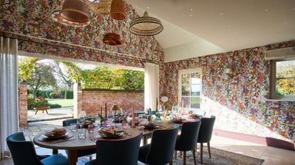 Image of an open room that leads to a back garden and has high slanted ceilings. There is a pink, floral-patterned wallpaper on the walls, and a dining table in the middle set for a dinner party.