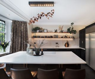 A stylish black and white kitchen with a large island hob. The space features leather bar stools, a sculptural copper chandelier, warm under-shelf lighting, and elegant decorative accents.