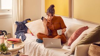 En kvinna sitter i en soffa i ett vardagsrum och dricker kaffe och kollar på en HP Chromebook som står placerad framför henne i soffan.