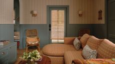 a cozy living room in a guest cottage with tiny floral wallpaper, denim blue half height paneling and trim, a patterned rust colored sofa with curved chaise and vintage coffee table