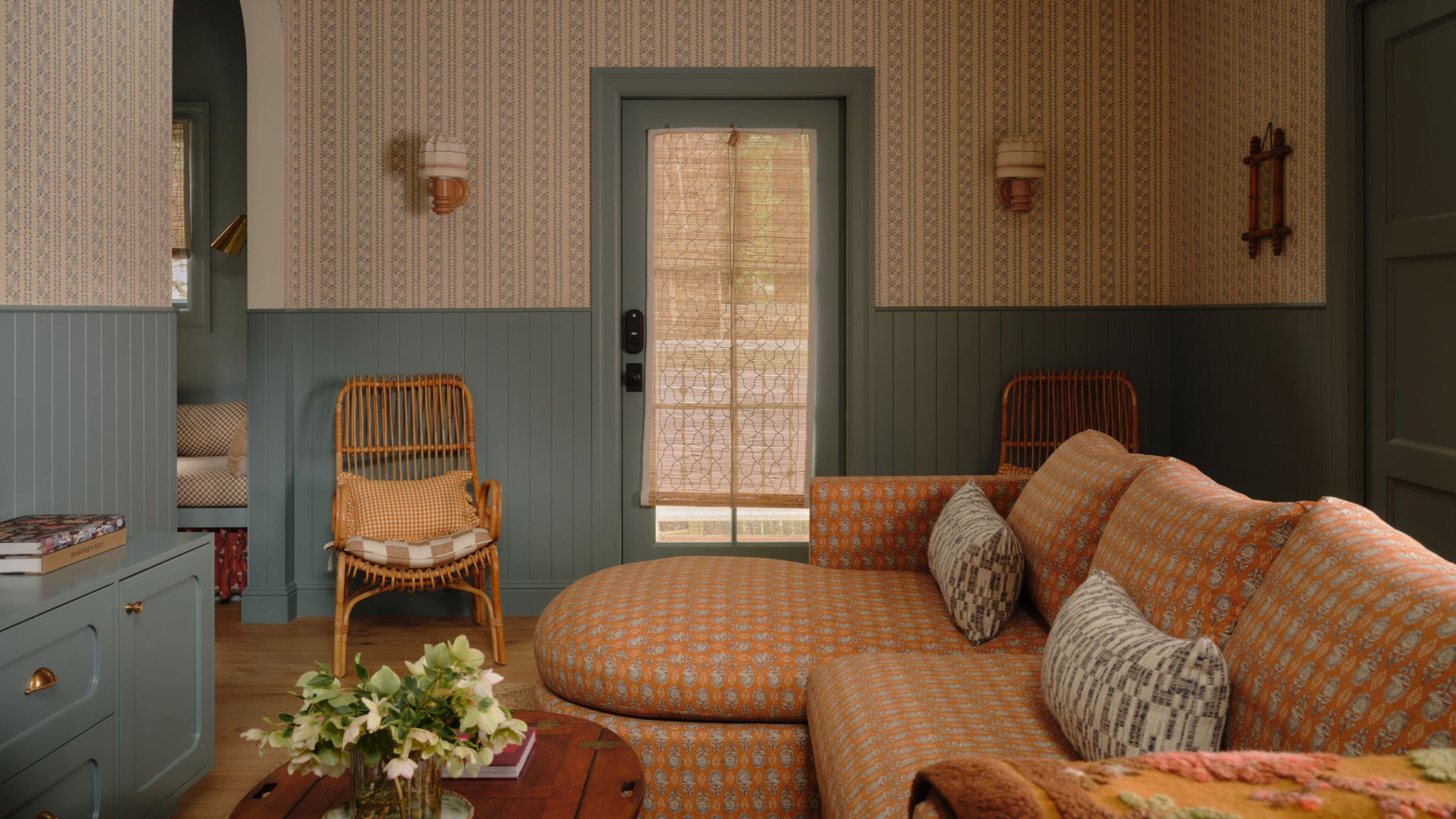 a cozy living room in a guest cottage with tiny floral wallpaper, denim blue half height paneling and trim, a patterned rust colored sofa with curved chaise and vintage coffee table