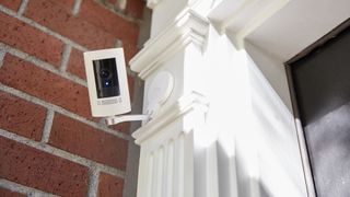 A white home camera with the recording light on mounted on a brick wall above a door painted white