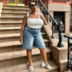 aniyah wears long jean shorts and white tank top and white sandals while standing at the bottom of a stoop