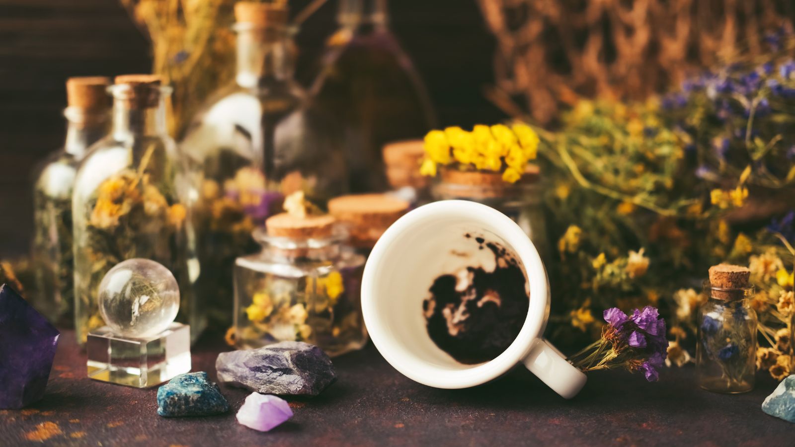 Composition of flipped over white cup with coffee grounds, colorful stones, bottles with dried flowers, oil and tincture, toy glass ball, dry herbs and a net.