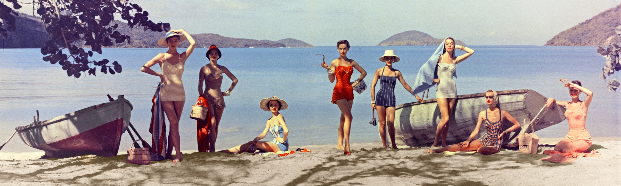 Nine women in 1950s swimsuits and sun hats pose along a sandy beach between two wooden rowing boats, with calm blue water and hilly islands behind them.