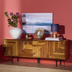 A wooden TV stand with indoor plants, ornaments, and lights on top and a wall picture above from Wayfair on a pink background