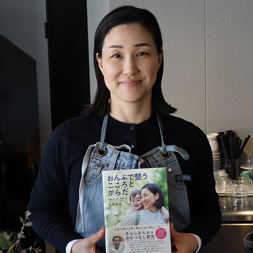 a smiling woman wearing an apron in a cafe holds up a book with a smiling mother and baby on the cover