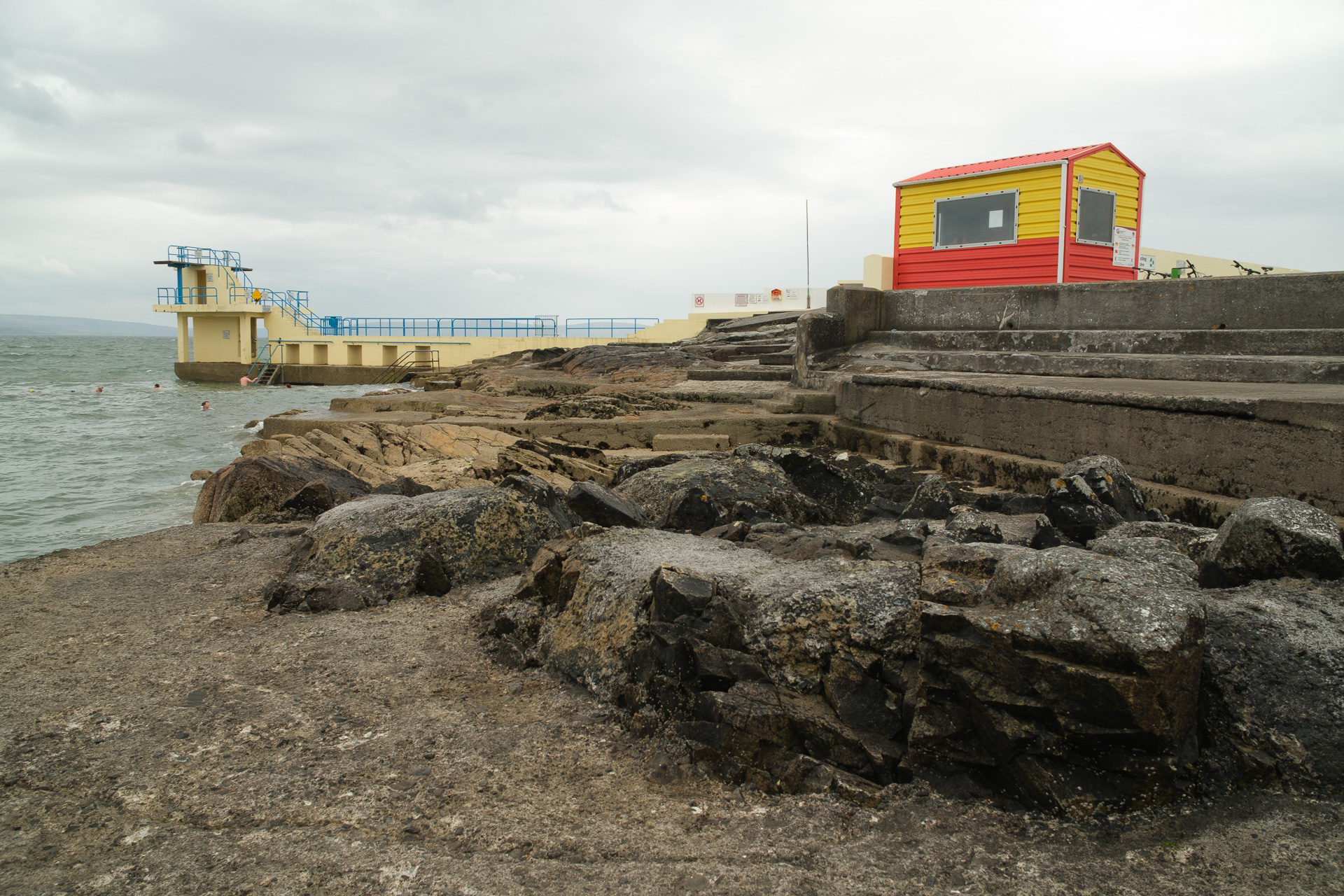 A rocky coastal swimming area with concrete terraces, a red and yellow hut and a diving tower under an overcast sky.