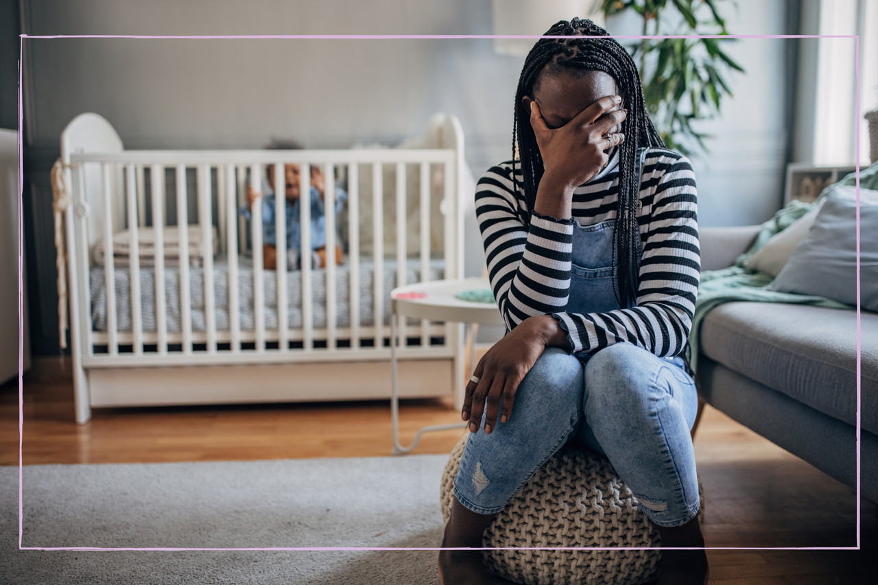 Exhausted mother sitting at home, her crying baby boy is in a crib behind her.