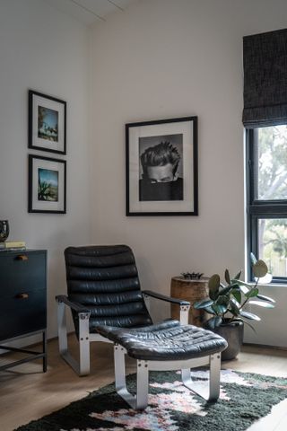 Corner of a room that has a black, modern lounge chair and a plant in the corner beside it. There are three pictures hanging on the wall and part of a window in view.