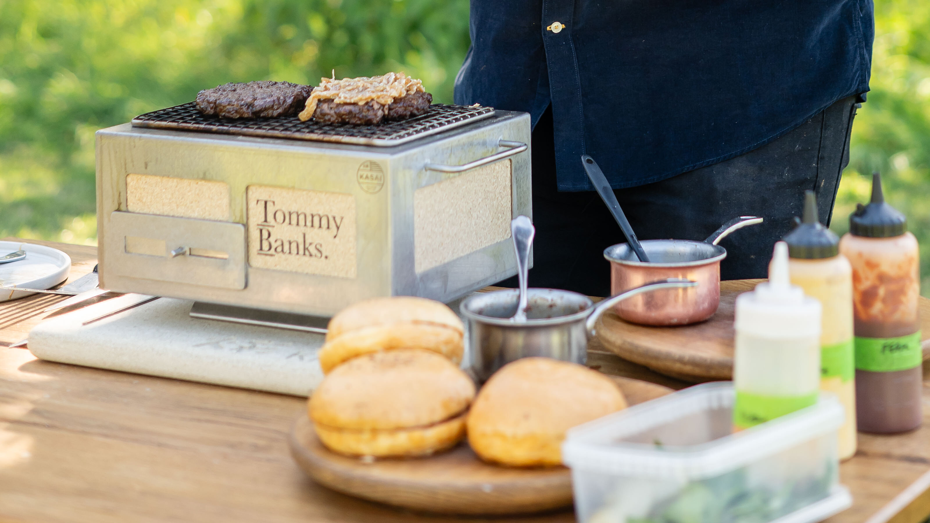 Close up of a steel Japanese style grill on a wooden outdoor table with burger buns and sauces 