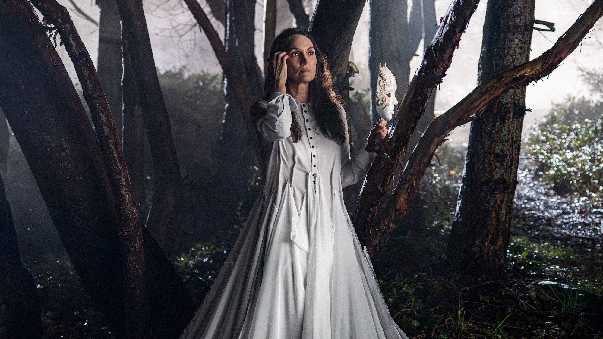 Carrie-Anne Moss' Katherine wearing a long off-white dress and holding a mask on a stick while standing in th ewoods in Yaga