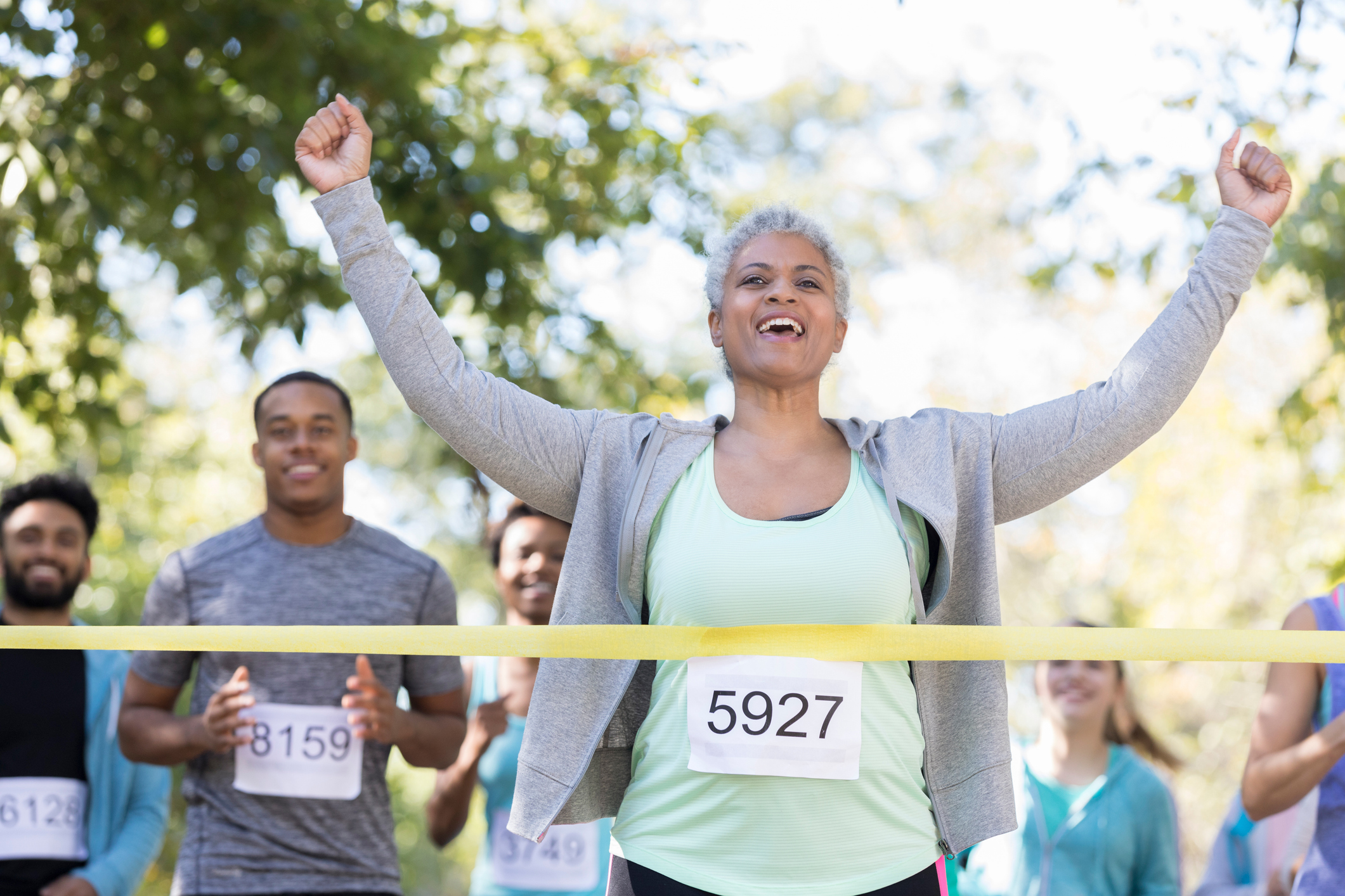 An ecstatic senior woman raises two fists in victory as she crosses the finish line of a charity race first. Other runners smile behind her.