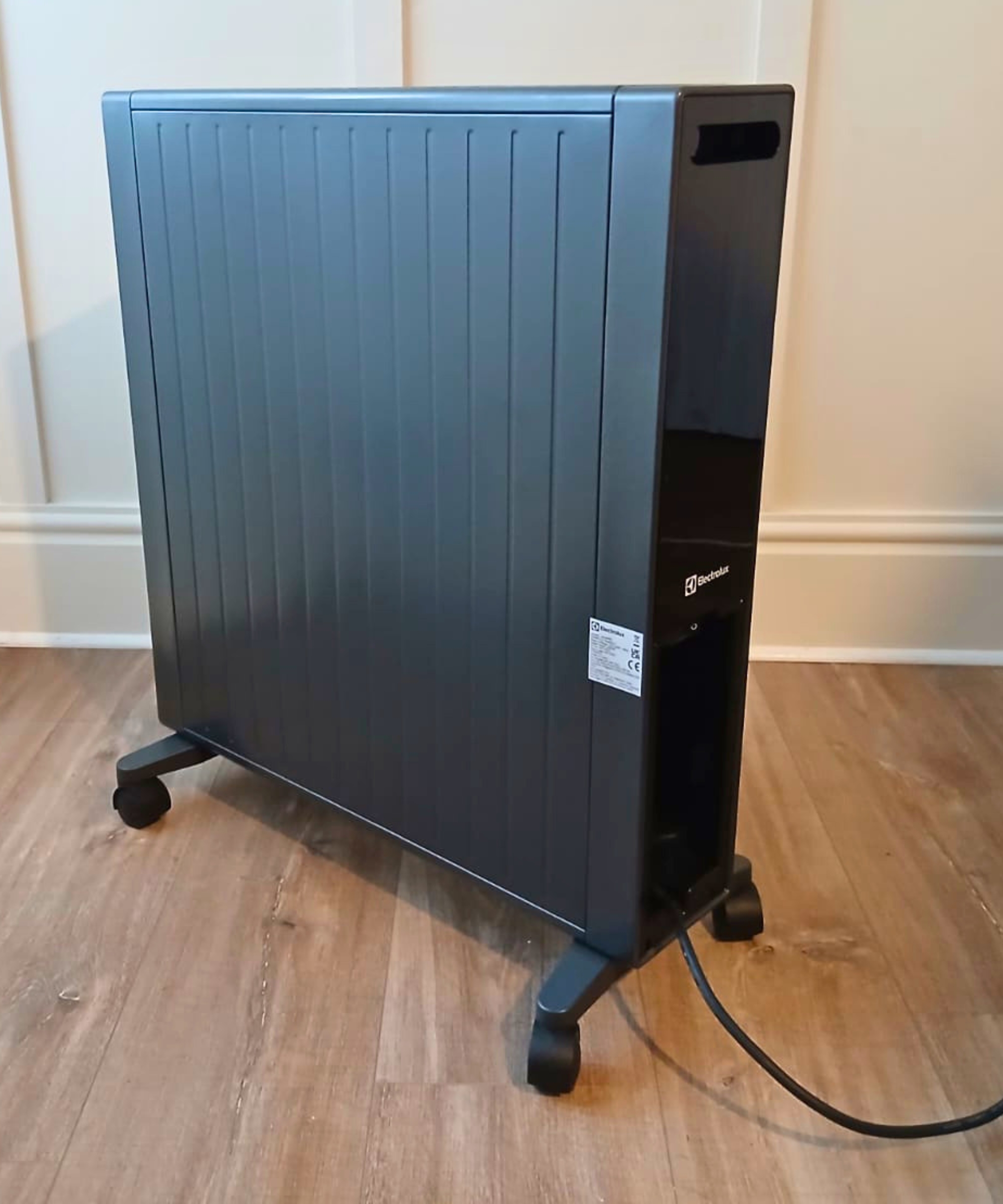 Dark grey Electrolux portable heater on castors, plugged in on a wooden floor.