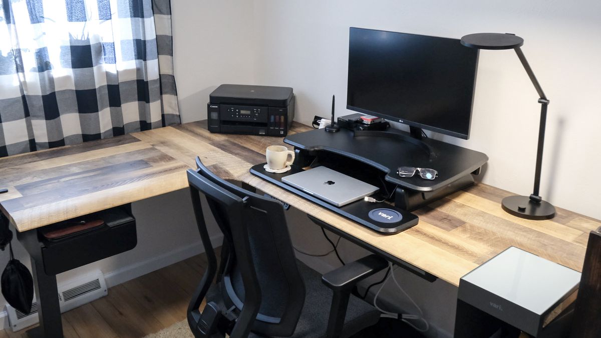 A side view of the Vari L-Shape Electric Standing Desk with a monitor, laptop, printer and desk lamp