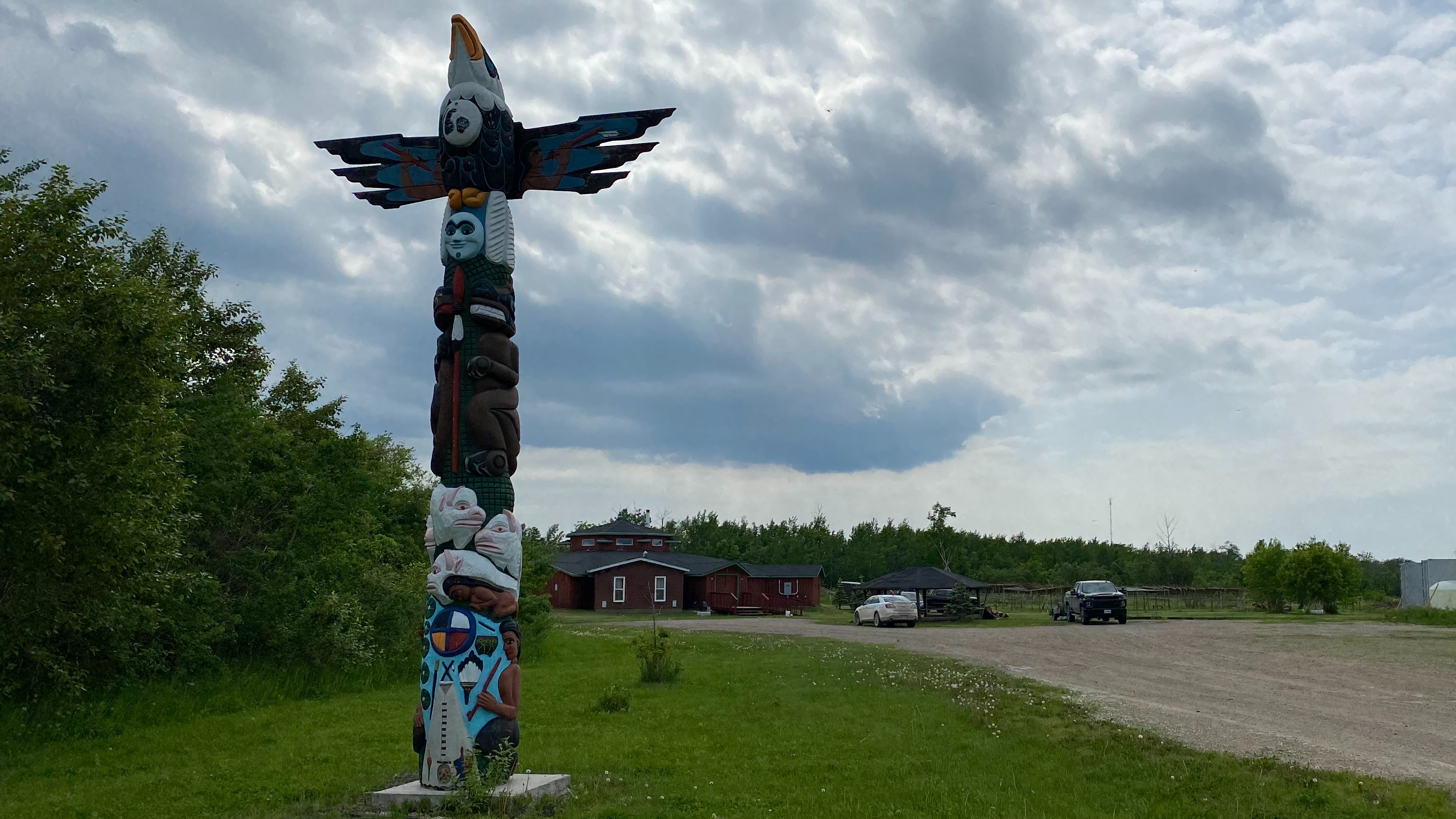 totem pole in the foreground of a picture with cloudy sky in behind. far in the back is a lodge and cars, with a road leading to it. trees surround the scene