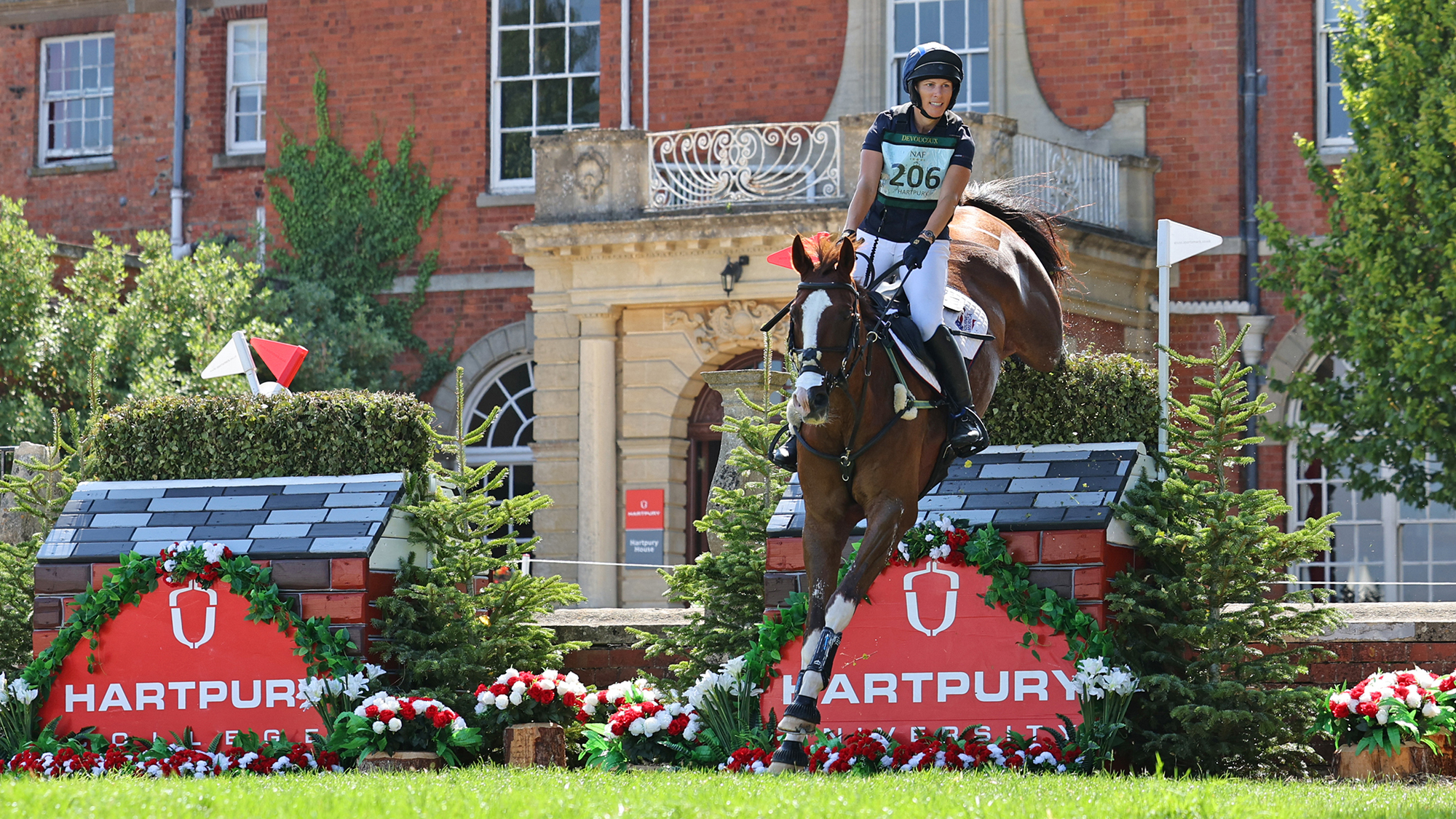 Zara Tindall jumps a skinny Hartpury-branded house on her chestnut horse Class Affair with the red brick of Hartpury College in the background