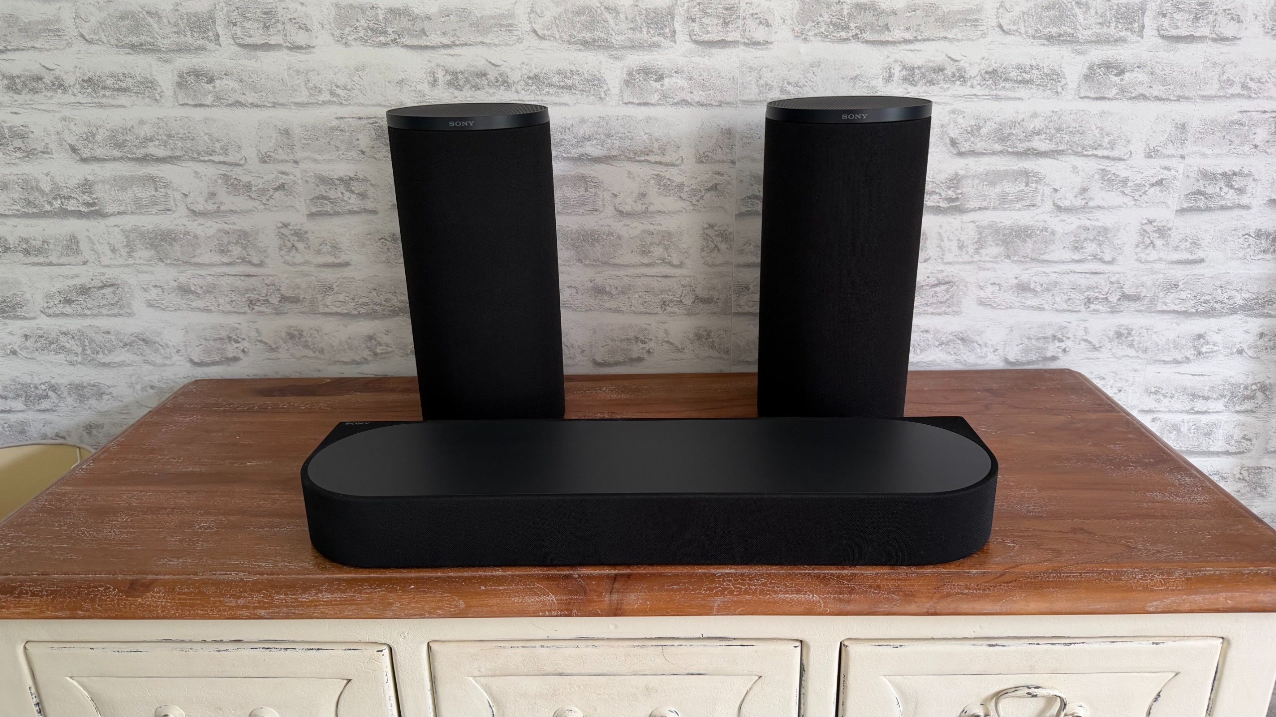 The Sony Bravia Theatre Trio sits on a wooden sideboard in front of a white brick wall.